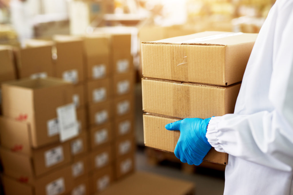 Close up view of a dedicated worker carrying a stack of duck taped brown boxes in factory storage room while wearing sterile cloths and rubber gloves.